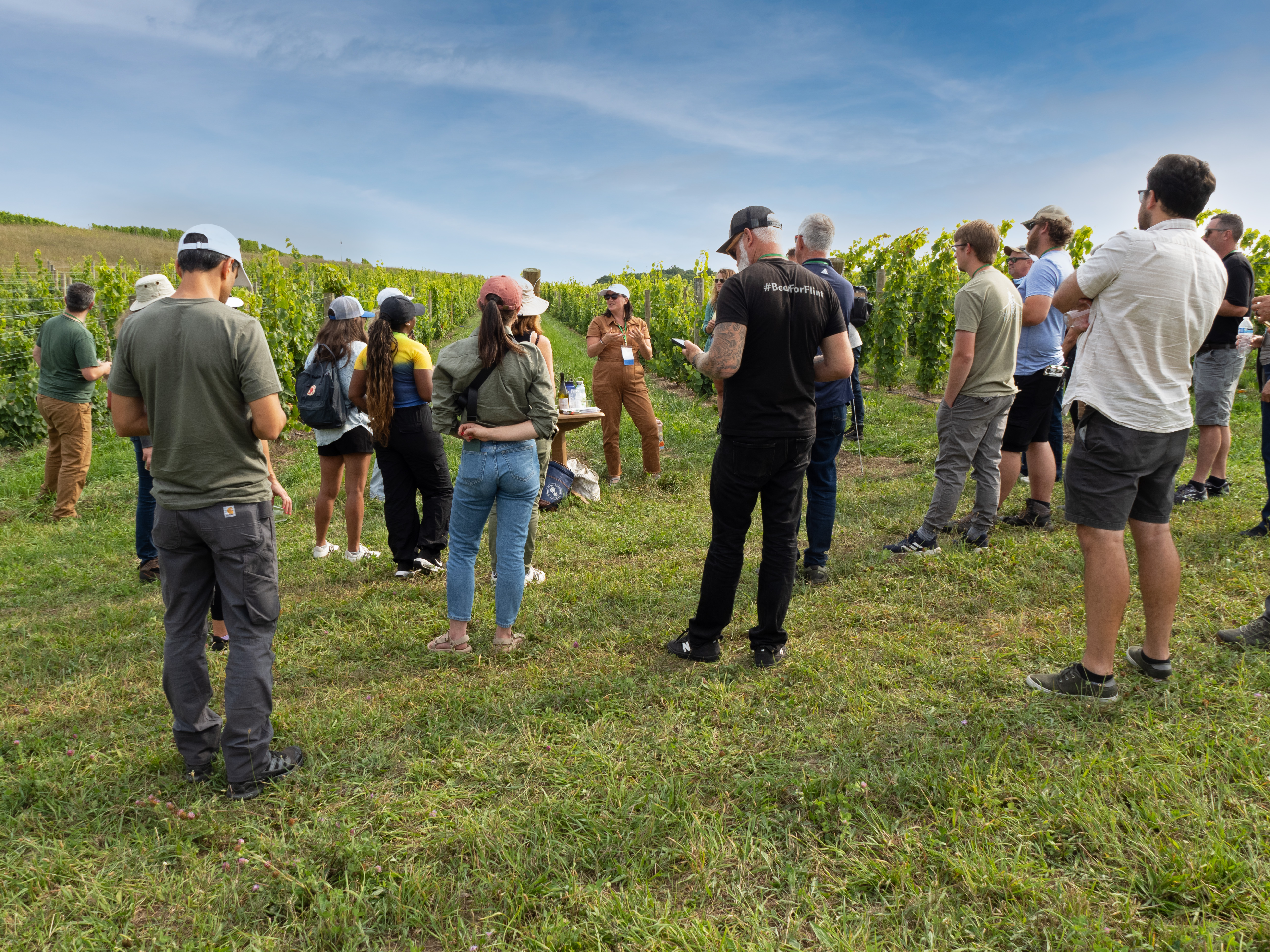 Attendees of Dirt to Glass Conference standing outside in a grape vineyard listening to a presenter talk to them.